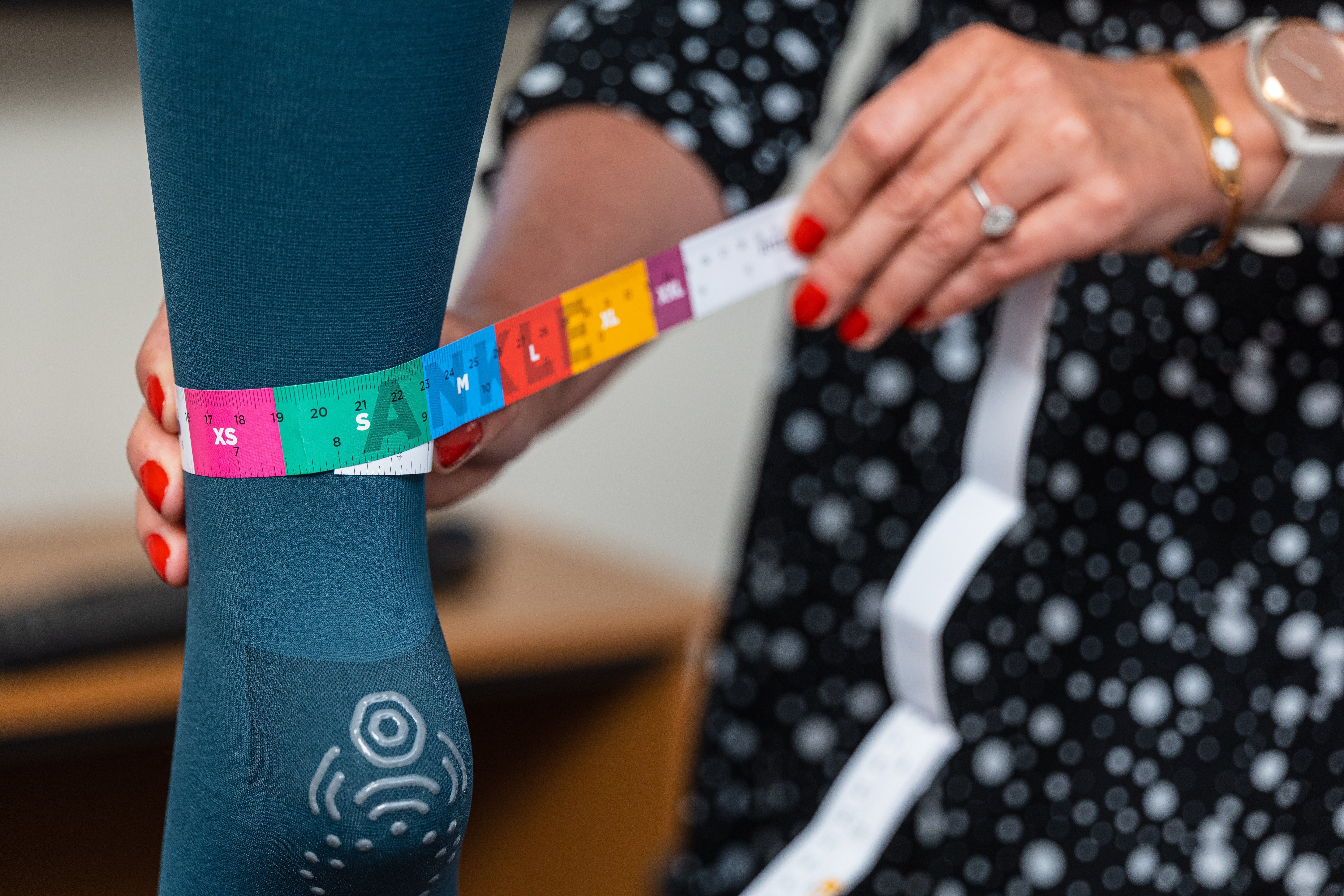 A close-up of a person using a color-coded measuring tape to measure the ankle circumference over a teal compression sock for accurate sizing.