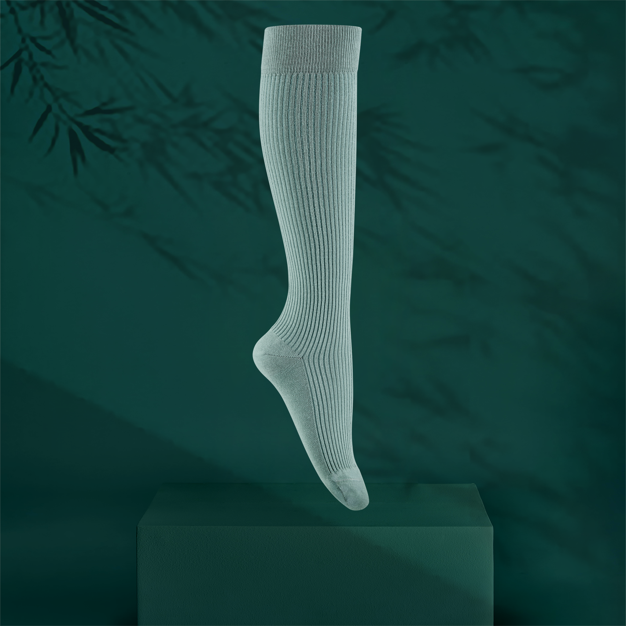 A single ribbed knee-high sock in a pale eucalyptus green colour, shown floating above a dark green pedestal against a dark teal background with soft bamboo leaf shadows.