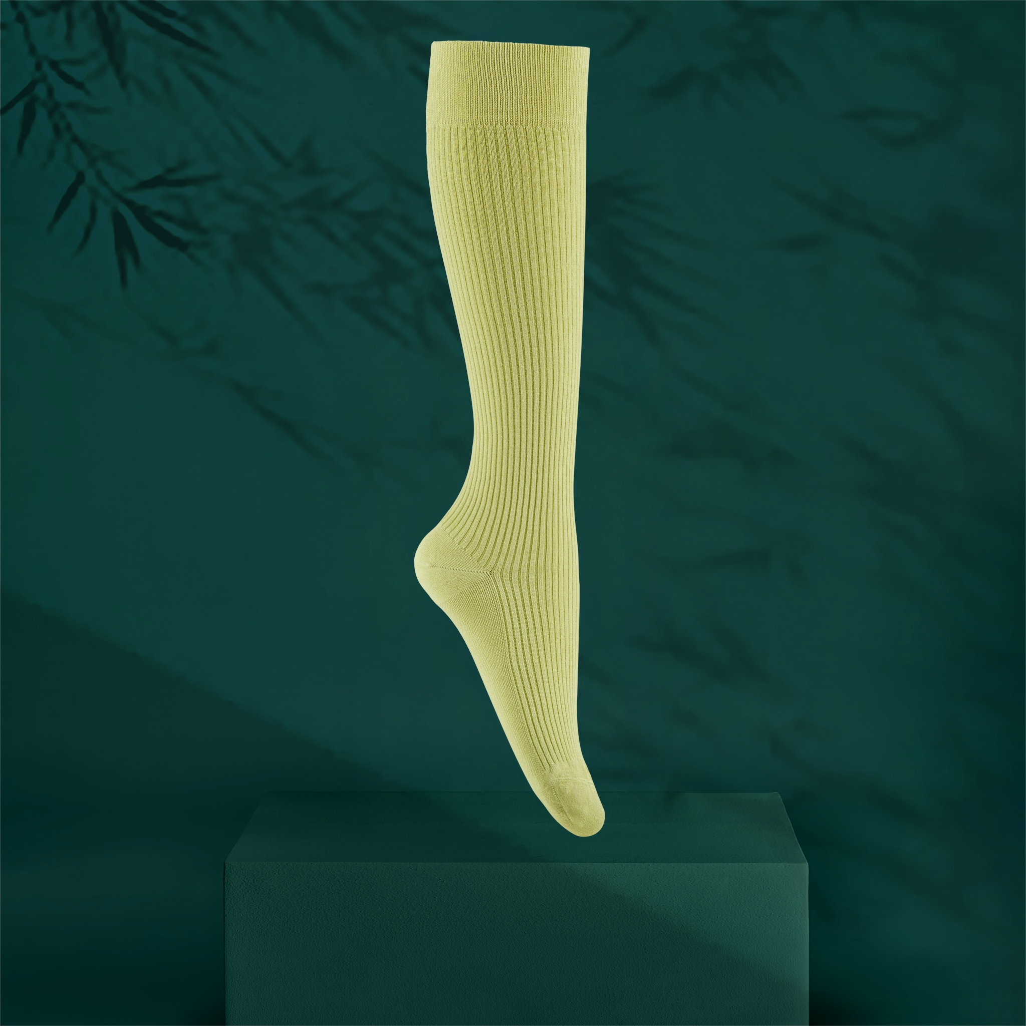 A single ribbed knee-high sock in a bright lemongrass green colour, shown floating above a dark green pedestal against a dark teal background with soft bamboo leaf shadows.