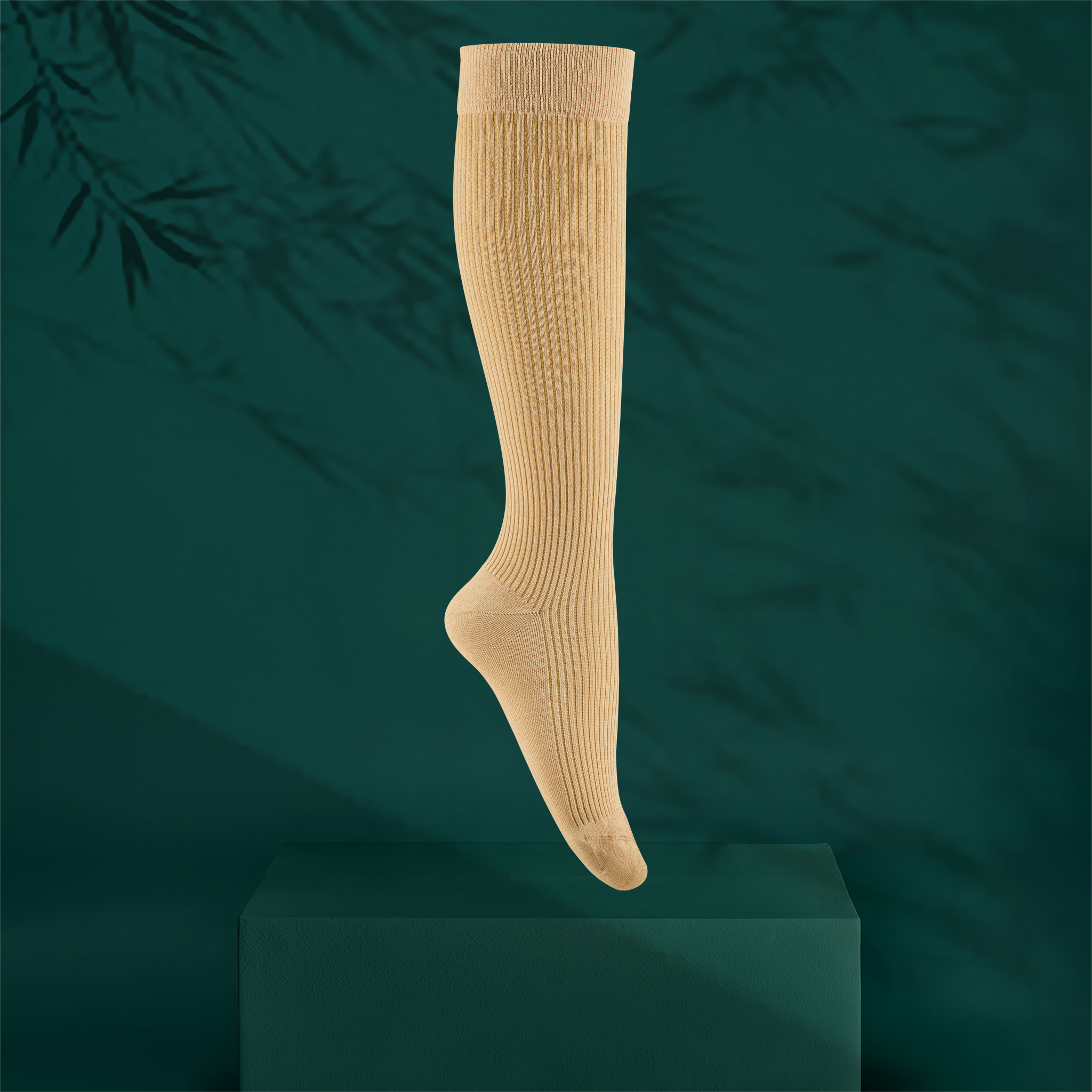 A single ribbed knee-high sock in a pale maple wood orange colour, shown floating above a dark green pedestal against a dark teal background with soft bamboo leaf shadows.