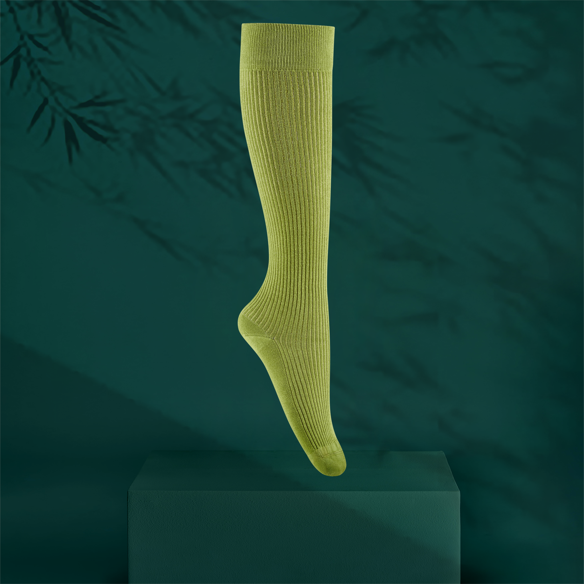 A single ribbed knee-high sock in an olive green colour, shown floating above a dark green pedestal against a dark teal background with soft bamboo leaf shadows.