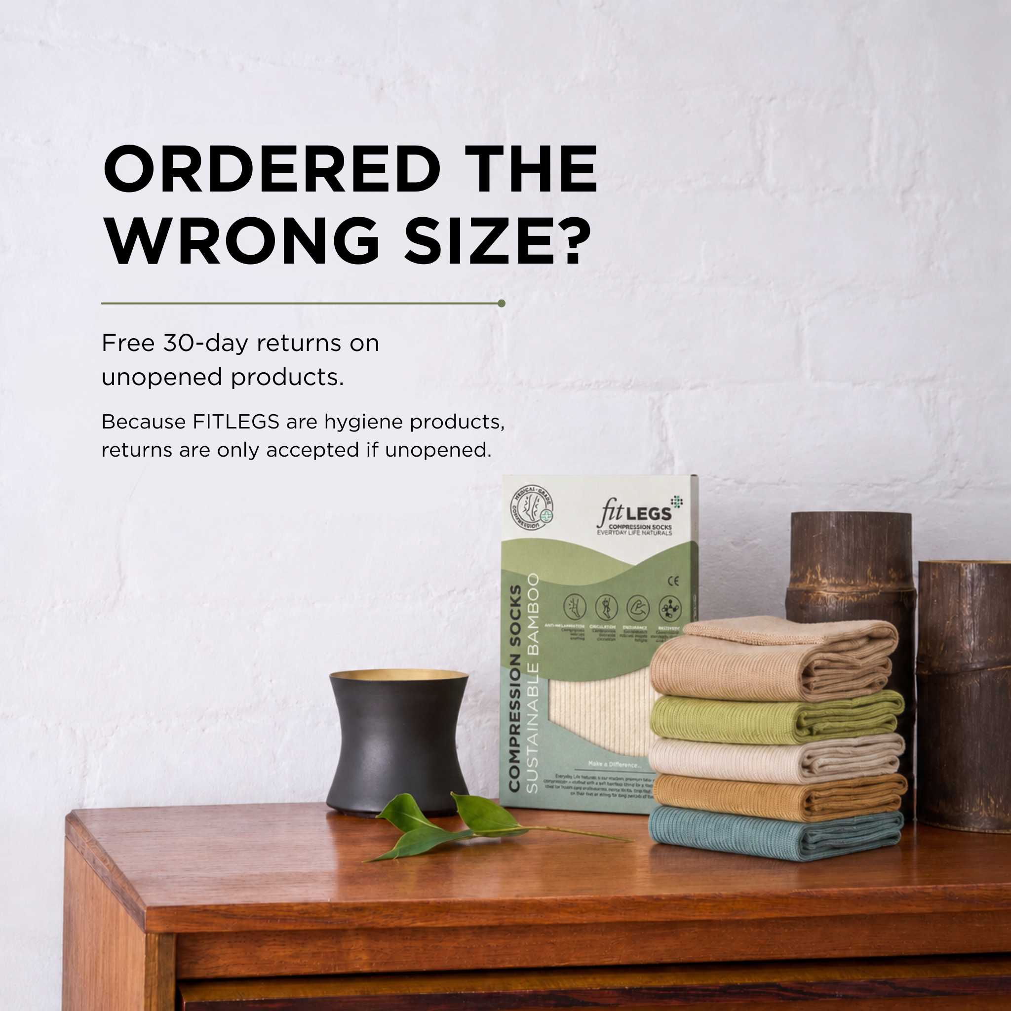 A box of FITLEGS Compression Socks stands on a wooden table next to a stack of folded socks in beige, green, tan, and blue green, with a black candle and green leaves. Text on the wall reads 'ORDERED THE WRONG SIZE?' with a note about free 30-day returns on unopened products.